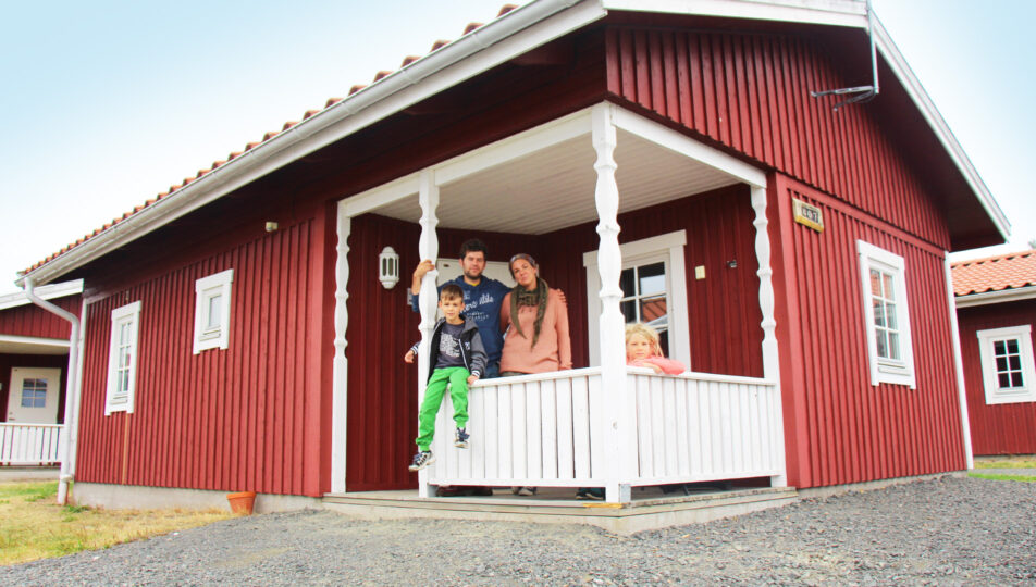 Röd stuga på Sunne camping med familj på farstukvisten, två vuxna och två barn.